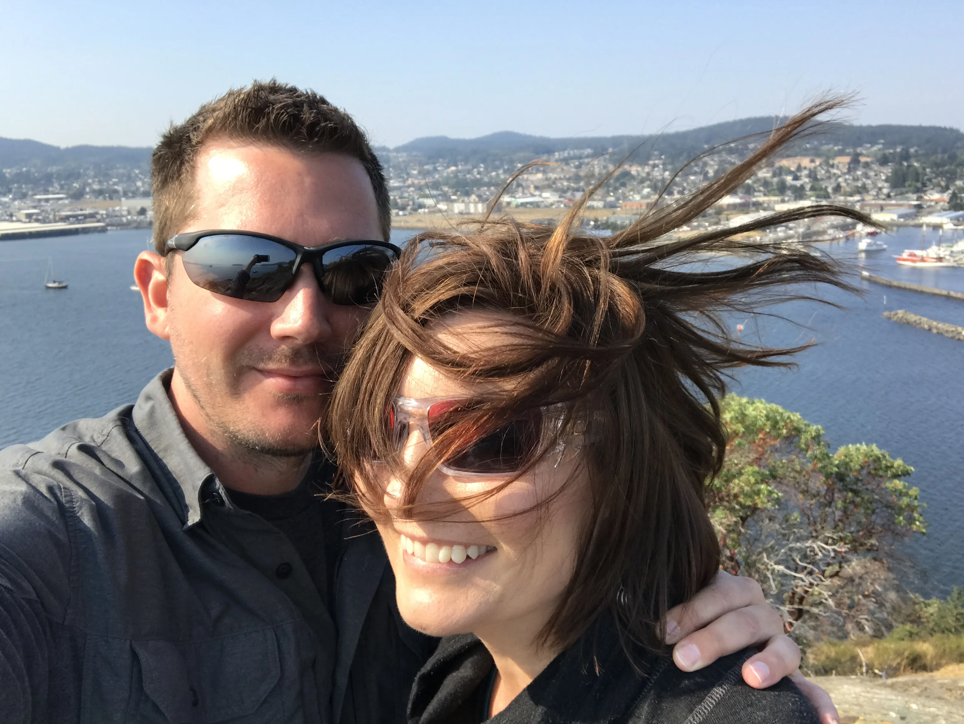 Stacy and Brandon stand together above Anacortes as wind blows Stacy’s hair across her face.