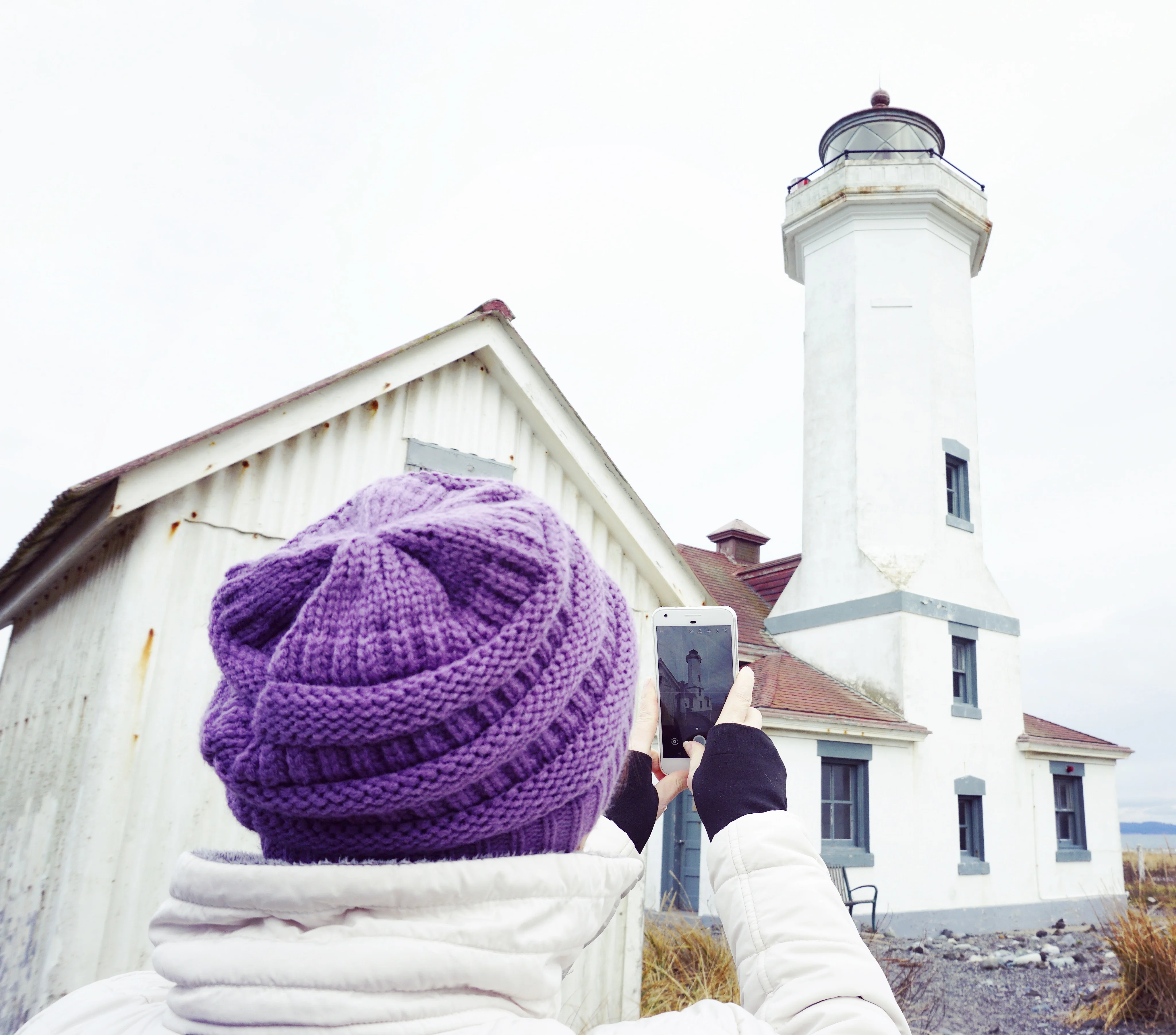 Stacy holds up her phone to photograph Point Wilson Lighthouse, visible both ahead of her and on the phone screen.
